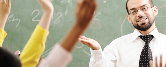 A teacher standing in front of a class with students raising their hands